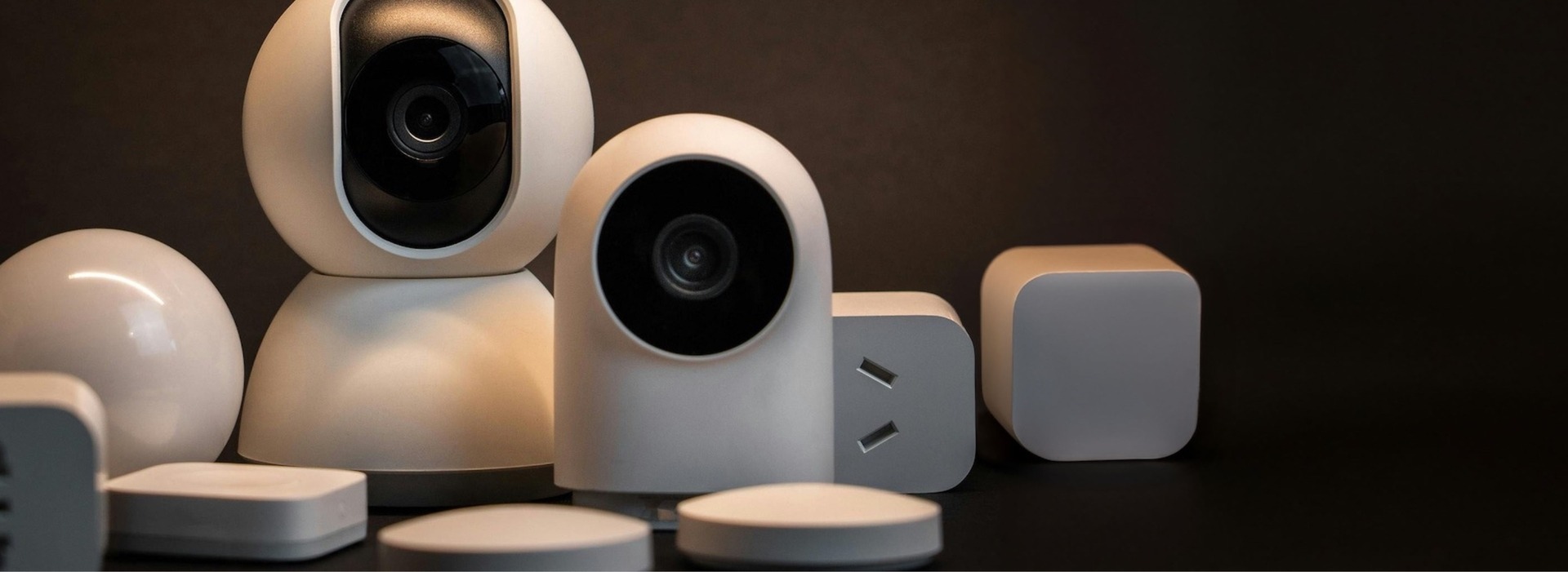 A collection of modern smart home devices on a dark background, including two white security cameras with lenses, power adapters with USB ports, and various small white sensors and hubs, arranged neatly together.