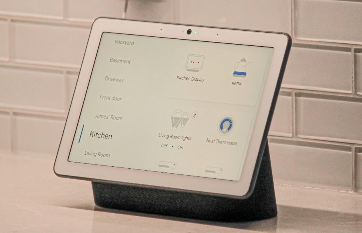 A smart home display device on a kitchen countertop with a white tiled backsplash, showing a menu on its screen with options for controlling various home areas like the kitchen, living room, and front door, including settings for lights and a Nest Thermostat.